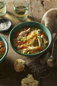 two bowls of chicken noodle soup on a wooden table with bread and water glasses