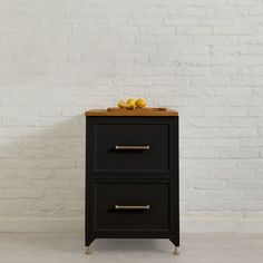 a black cabinet with two drawers next to a white brick wall and yellow fruit on top