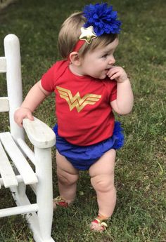 a baby girl wearing a red shirt and blue shorts standing next to a white chair