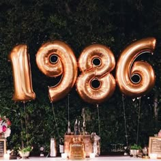 a table topped with lots of gold balloons and flowers next to a wall covered in greenery