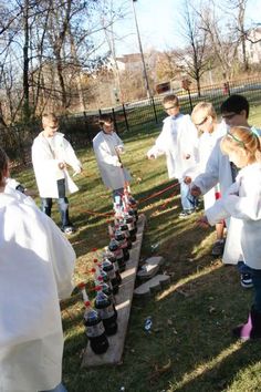 several children in lab coats are playing with cups and bottles on a wooden table outside