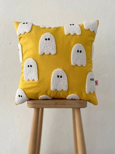 a yellow pillow with white ghost faces on it, sitting on a wooden stool in front of a white wall