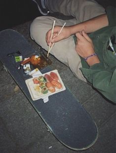 a person sitting on the ground with food and chopsticks in their hands next to a skateboard