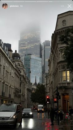 cars driving down a city street with tall buildings in the background and foggy skies