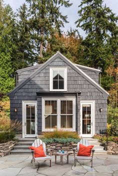 two chairs and a table in front of a small gray house with trees around it