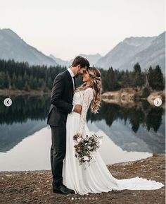 a bride and groom standing next to each other in front of a lake surrounded by mountains