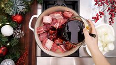 a person pouring sauce into a pot filled with meat and vegetables next to christmas decorations