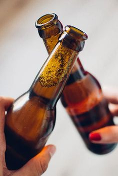 two brown beer bottles being held up by someone's hand with red nail polish on their nails