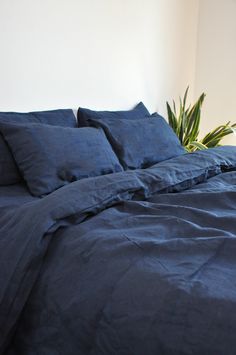 an unmade bed with blue linens and pillows in a white walled room next to a potted plant