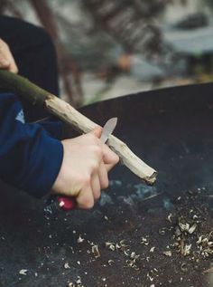 a person holding a wooden stick over an open fire pit with wood shavings on it