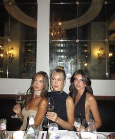 three women sitting at a table with wine glasses in front of them, posing for the camera