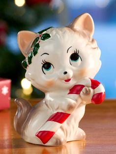 a white ceramic cat figurine holding a candy cane on a table next to a christmas tree