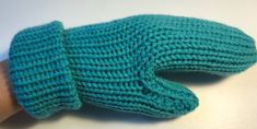 a hand knitted green mitten sitting on top of a white table next to a person's arm