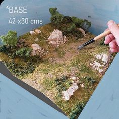 a hand holding a paintbrush in front of a miniature landscape