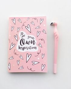 a pink notebook sitting next to a pen on top of a white surface with hearts and the words be your own inspiration