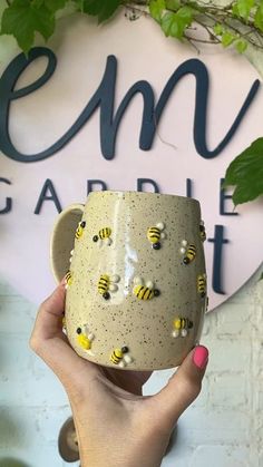 a hand holding a coffee mug with bees on it in front of a sign that says emu garden