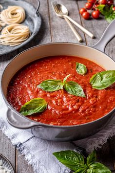 a pan filled with sauce and basil leaves on top of a table next to pasta