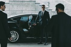 a man in a suit and sunglasses getting out of a car with another man standing next to him