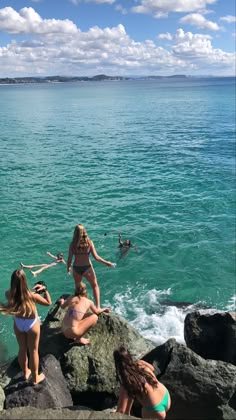 three women in bikinis are sitting on rocks near the water and one is standing