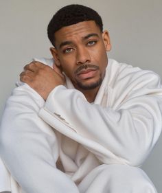 a man with his arms crossed sitting on the floor wearing a white sweatshirt and sweatpants