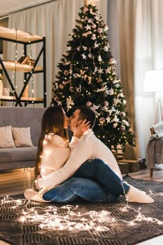 a man and woman are sitting on the floor kissing in front of a christmas tree