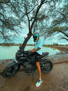 a man sitting on top of a black motorcycle next to a tree and body of water