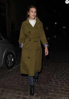 a woman in a trench coat is walking down the street at night with her hand on her hip