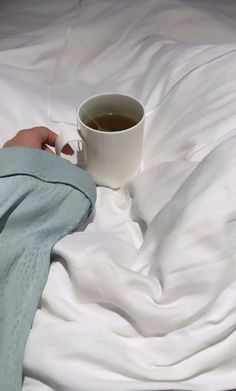 a person's hand holding a cup of tea on top of a white bed