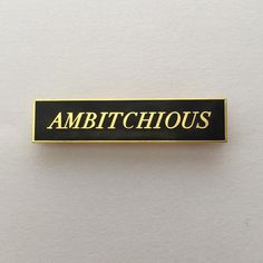 a black and gold pin with the words ambitioushouse on it's back side