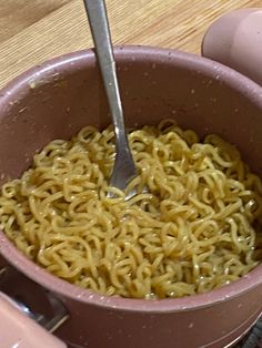 someone is holding a spoon over some noodles in a pink bowl on top of a wooden table