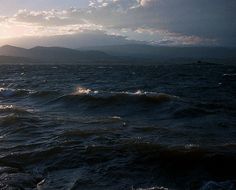 the sun shines through clouds over the ocean as waves crash on the water and mountains in the distance