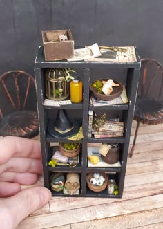 a hand holding a miniature shelf with many items in it on top of a wooden table