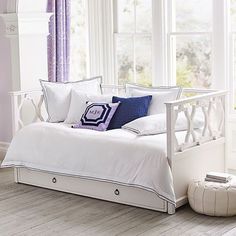 a white day bed with purple and blue pillows on it in front of two windows