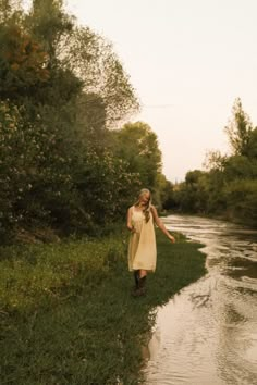 a woman in a dress is walking on the side of a river with her arms outstretched