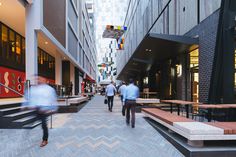 people are walking down the sidewalk in an urban area with tables and benches on either side