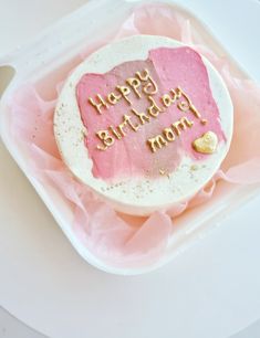 a birthday cake with pink frosting and gold writing on it sitting in a plastic container