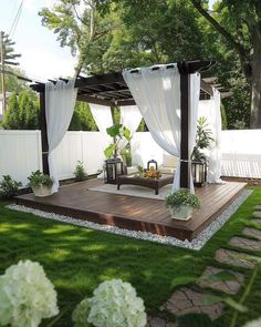 a wooden deck with white curtains and flowers in the grass next to a fenced area