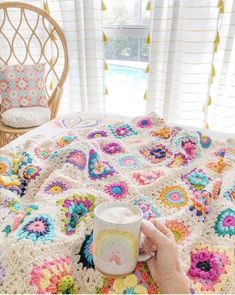 a woman is holding a coffee cup in her hand while sitting on a bed with a crocheted blanket