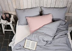 an open book on a bed with grey sheets and pillows, next to a white chair