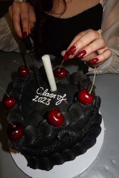 a woman is cutting into a black cake with cherries on the top and candles in the middle