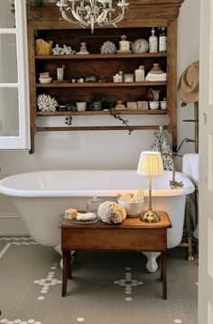 a white bath tub sitting under a chandelier next to a wooden shelf filled with bottles