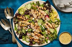 a salad with chicken, cucumbers and dressing on a blue tablecloth next to utensils