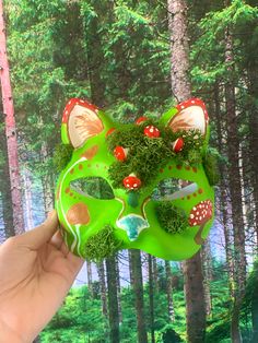 a hand holding up a green mask with mushrooms on it in front of a forest