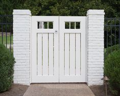 an open white gate in front of a brick wall and shrubbery on the other side