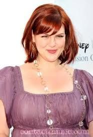 a woman with red hair wearing a purple dress and large earrings on her neck, posing for the camera