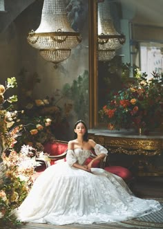 a woman in a white dress sitting on a red couch next to a mirror and chandelier