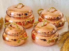 four copper pots with lids are sitting on a table next to dried grass and dry stalks