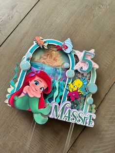 an ariel the mermaid birthday cake topper on a wooden table with disney characters and numbers