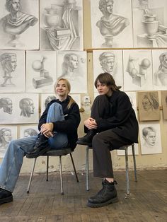 two women sitting on chairs in front of wall with drawings and busturines behind them