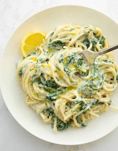 a white plate topped with pasta covered in spinach and lemon wedges next to a fork